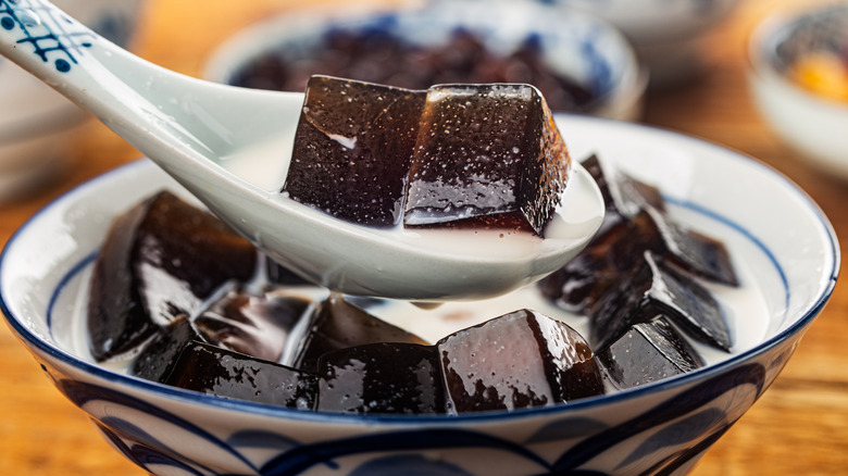 A bowl of grass jelly cubes in milk with a spoon lifting two pieces.