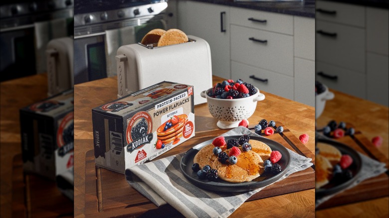 A box of Kodiak Power Flapjacks sits near a toaster with two pancakes and a prepared plate of pancakes.
