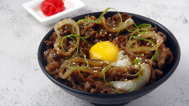 A bowl of gyudon toppen with an egg yolk sits on a table with condiments.
