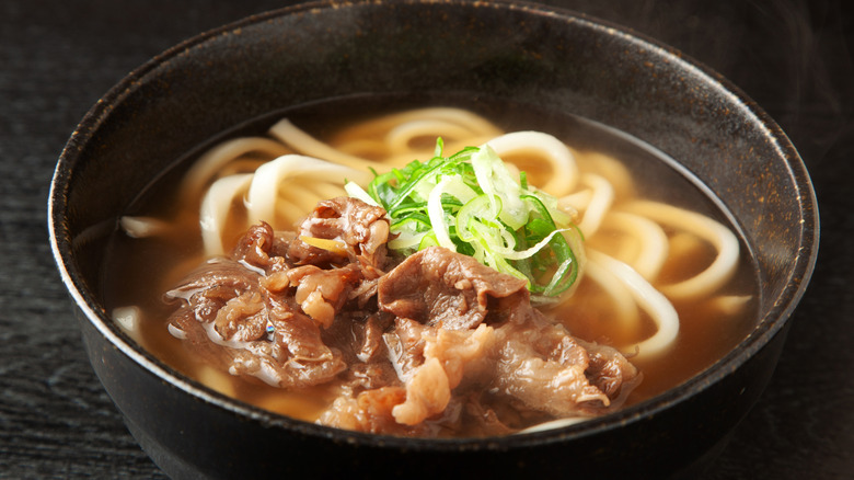 A bowl of udon noodles and beef in broth is on a table.
