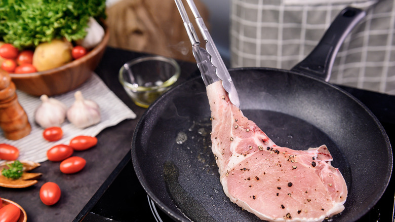 hands with tongs about to flip raw pork chop cooking in cast iron pan