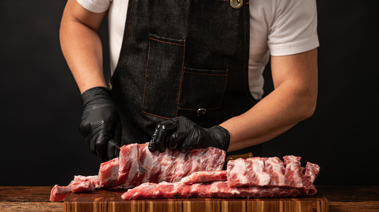 butcher with black apron and gloves cutting into rack of raw pork
