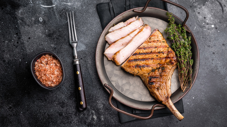 large pork chop sliced on a cast iron pan with thyme next to a fork and a bowl of pink salt