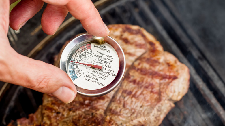 hand holding meat thermometer with temperature indicator above piece of meat on grill