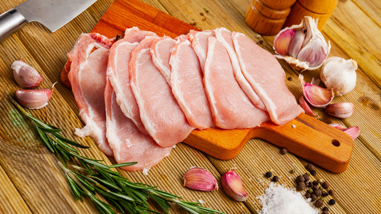 thin raw pork chops piled on top of cutting board surrounded by herbs, garlic, salt, and pepper