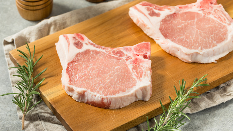 two raw pork chops with fat caps on cutting board with herbs