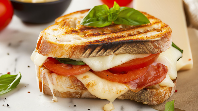 Basil, cured meat, and tomato topped grilled cheese sandwich overflowing on toasted bread