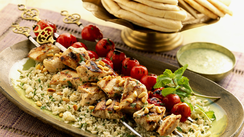 Skewers of chicken and tomatoes laid on a bed of rice in front of a platter of pitas