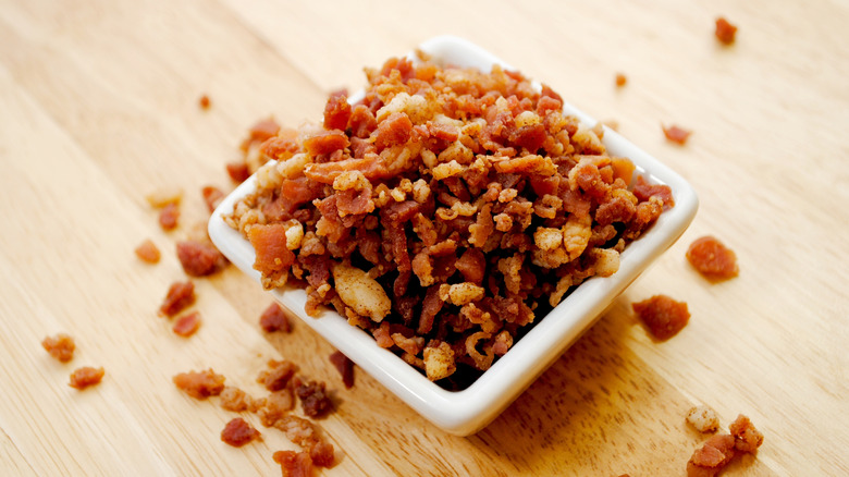 Crumbled bacon bits overflowing from a small dish onto a natural wood surface