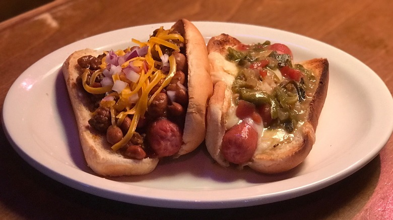 Chili cheese dog topped with red onion plated beside a hot dog smothered with chilis and cheese