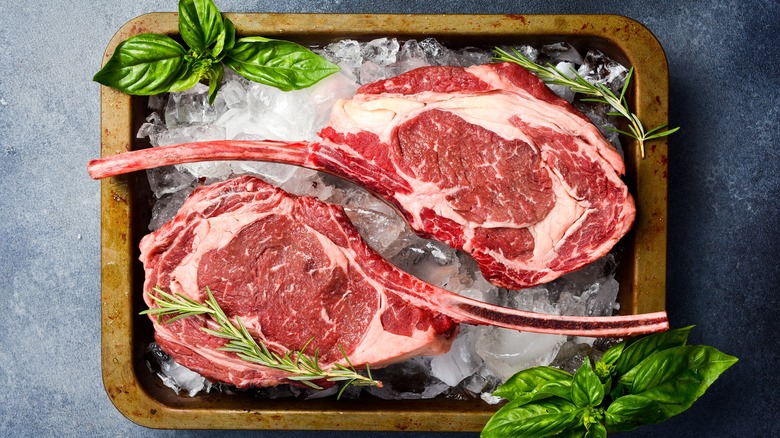 Two raw tomahawk steaks on ice with basil leaves and rosemary