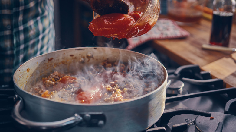 hand spooning tomatoes into a pot of bolognese cooking on the stove