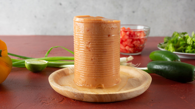 A molded can of refired beans standing on a wooden plate with fresh ingredients surrounding it