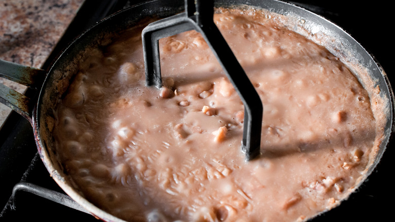Skillet of beans in liquid with a mashing utensil