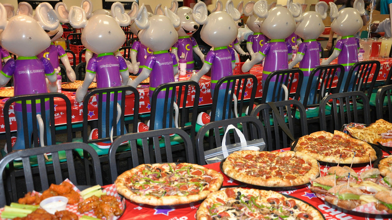 A table full of pizzas in front of a row of Chuck E. Cheese animatronics.