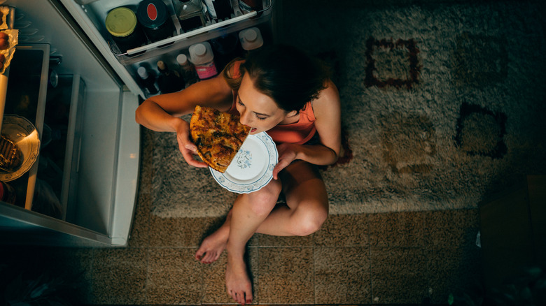 A woman sitting on the floor eating a slice of pizza in front of an open fridge.