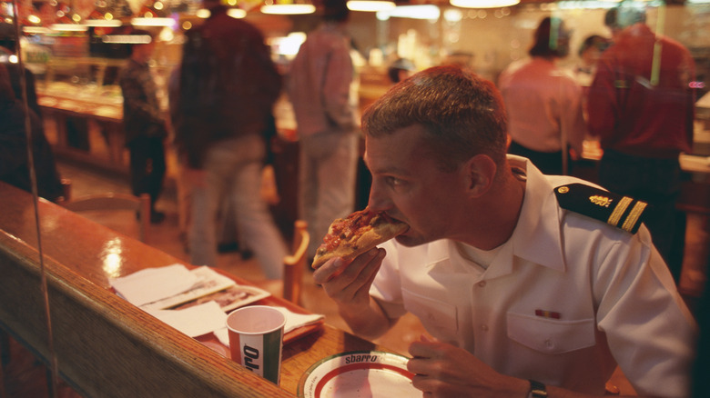 A pilot eats a slice of Sbarro pizza with diners lined up behind him.