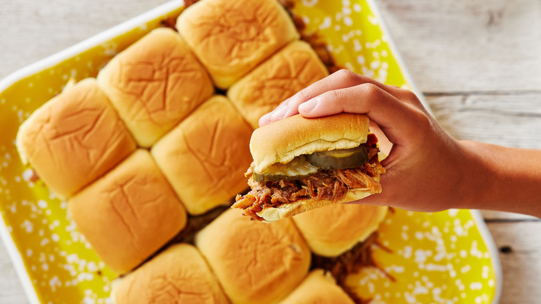 A hand holds a pulled pork slider above 11 others.