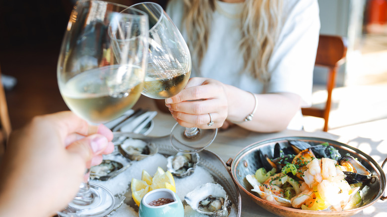 two hands clinking wine glasses above a table with oysters and a seafood dish