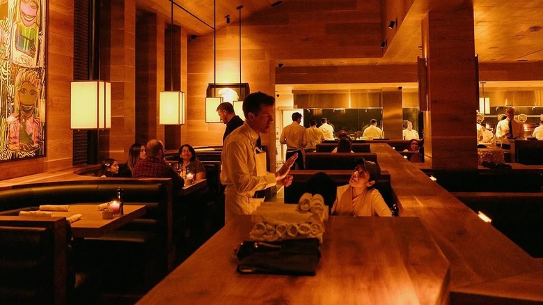 Interior of large wood-paneled restaurant showing servers and diners with kitchen staff in the background
