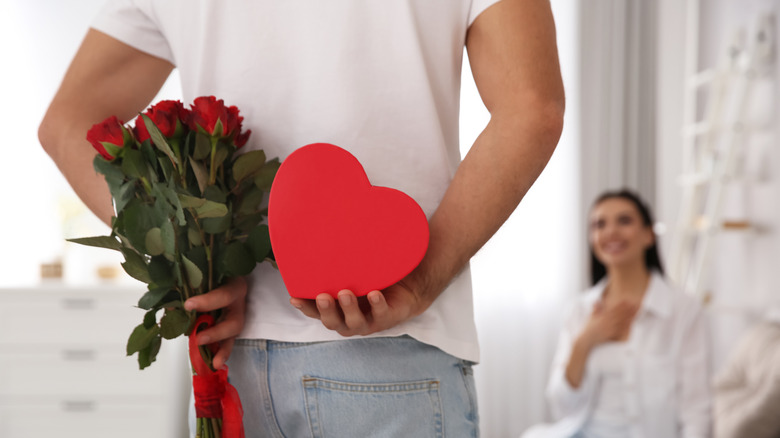 A romantic hides roses and a heart-shaped box behind their back, while a happy partner watches in the background