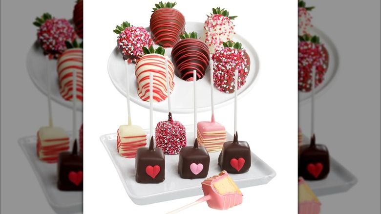 A plate of decorated chocolate-dipped strawberries sits next to a tray of chocolate-dipped cheesecake pops