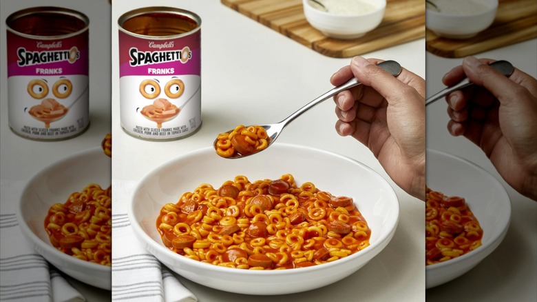 A hand takes a spoonful of SpaghettiOs with Franks from a white bowl, with an empty product can in the background