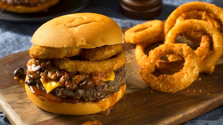 A burger topped with chili and onion rings sits on a wooden serving board next to a mound of onion rings