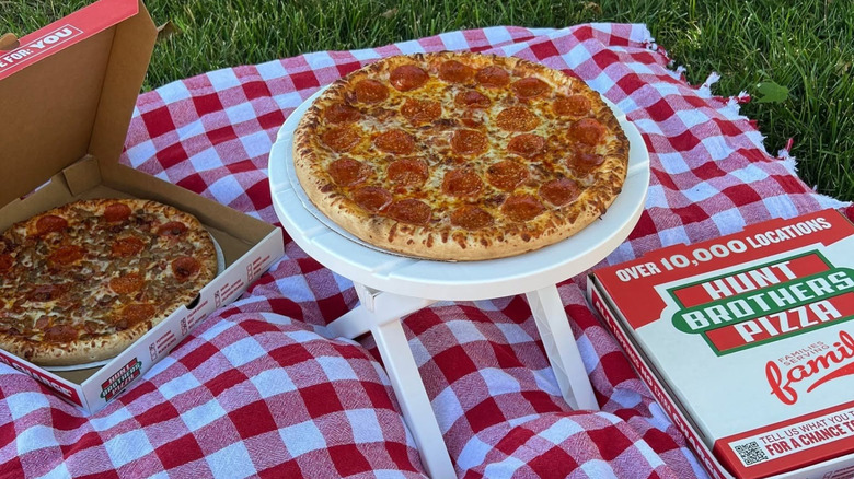 A Hunt Brothers pepperoni pizza sits on a small table at a picnic next to two pizza boxes.