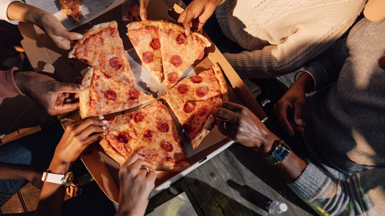 A group of friends all take slices of a pepperoni pizza out of its box.