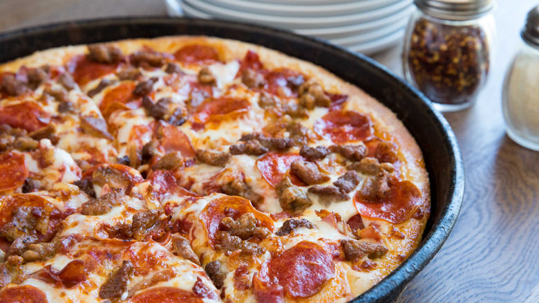 A Pizza Hut Meat Lovers pizza on a restaurant table next to a stack of plates and shakes of pepper flakes and parmesan