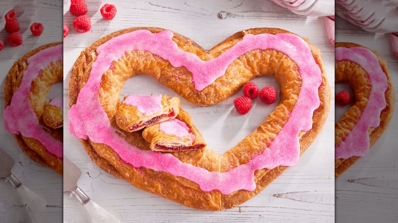 A heart-shaped kringle with pink icing and raspberries sitting nearby
