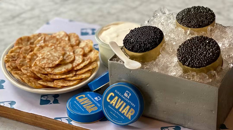 A mother of pearl caviar spoon balances on a box holding three open tins of caviar on ice next to the tin lids, a pot of crème fraiche, and a plate of Russian blinis.