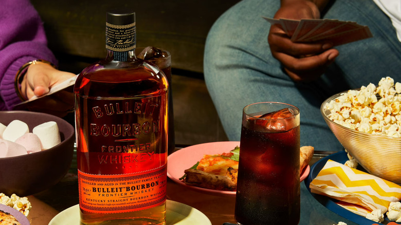 A bottle of Bulleit Bourbon Whiskey sits on a snack table with people playing cards in the background.