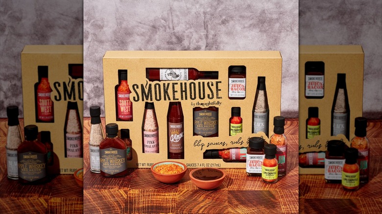 A Smokehouse by Thoughtfully Ultimate BBQ Gifts Sampler Set surrounded by bottles of rub and sauce and dishes containing spices.