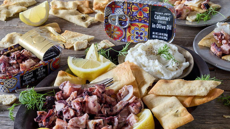 Tins of Trader Joe's Calamari Pieces in Olive Oil sit near a spread of pita chips, lemon wedges, and served calamari.