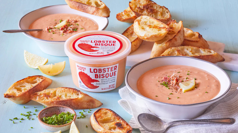 A container of Trader Joe's Lobster Bisque is surrounded by bowls of prepared soup and slices of bread.