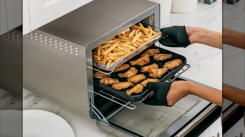 Hands wearing silicon oven mitts pull trays of wings and french fries from a Ninja Foodi 10-in-1 Smart Air Fryer Oven