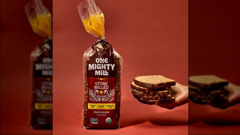 A hand holds bread slices next to a bag of One Mighty Mill Power Grains Bread.