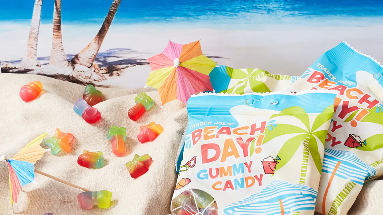 Two bags of Beach Day! Gummy Candy sit on sand-colored fabric against a beach background, with paper drink umbrellas and gummies sitting adjacent.