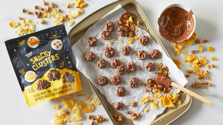 A bag of Trader Joe's Snacky Clusters sits next to a tray of clusters, crushed bits of salty snacks, and a bowl of chocolate