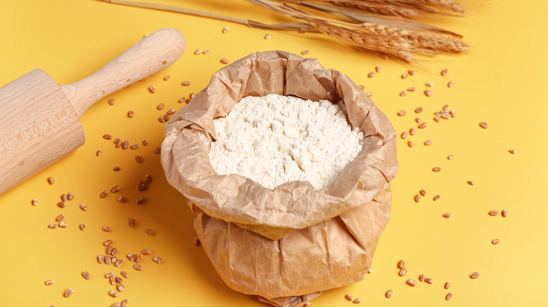 A sack of flour sits on a yellow surface next to a rolling pin and stalks of wheat
