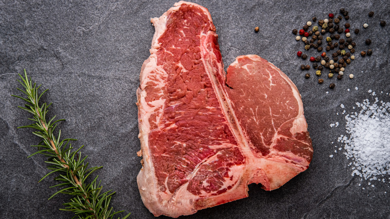 A raw porterhouse steak on a gray slab surrounded by salt, pepper, and herbs