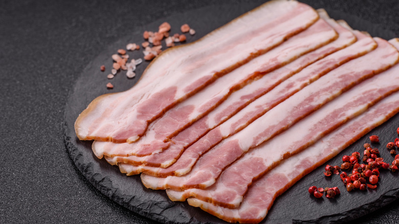 Raw bacon slabs on a black serving stone alongside seasonings