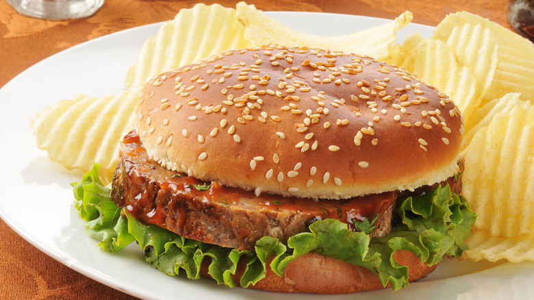 Meatloaf sandwich with lettuce on a round seeded bun with potato chips on the side