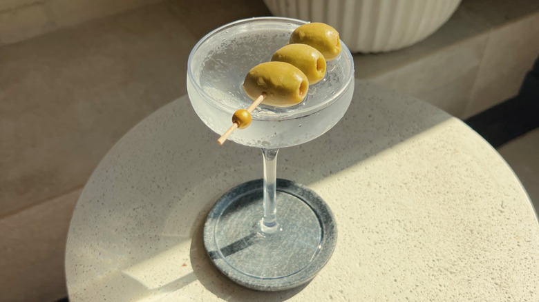 a downward view of a chilled dirty martini, garnished with three olives on a white, patio table