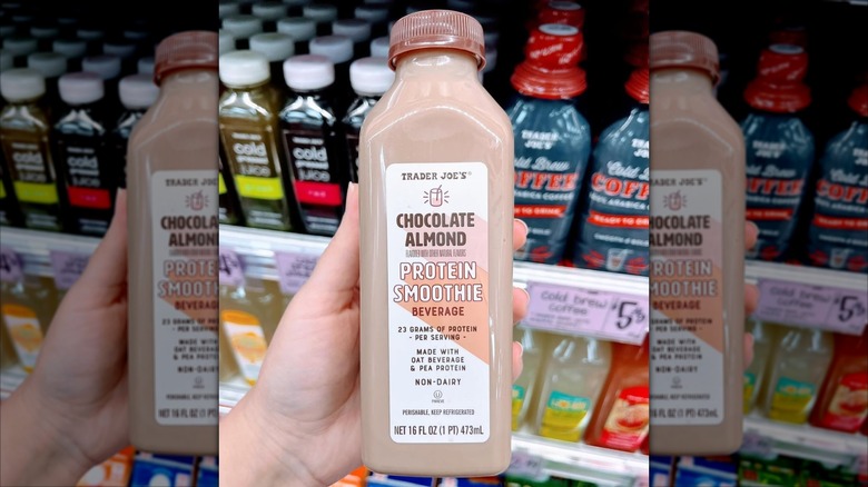 A hand holds up a Trader Joe's Chocolate Almond Protein Smoothie Beverage in front of a refrigerator case.