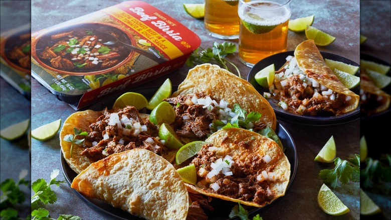 A package of Trader Joe's Beef Birria sits next to plates of tacos, glasses of drinks, and lime wedges
