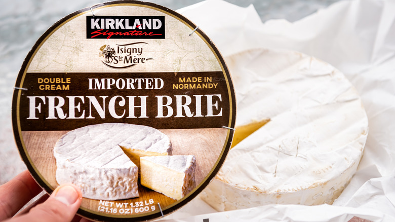 A person is holding a package of Kirkland Signature Imported Double Crème French Brie Cheese.