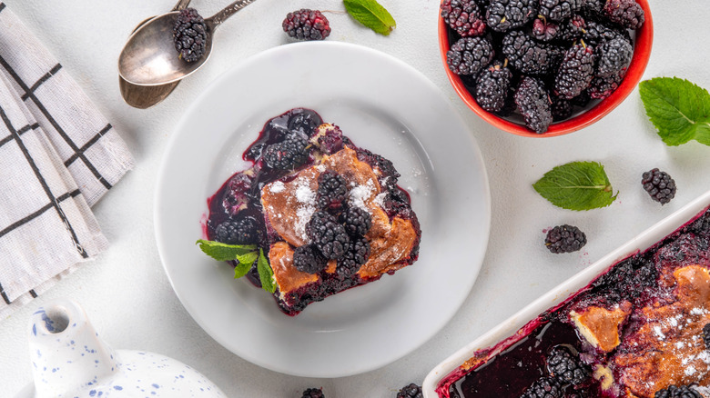 A piece of blackberry cobbler on a plate next to a bowl of blackberries and a baking tray of cobbler